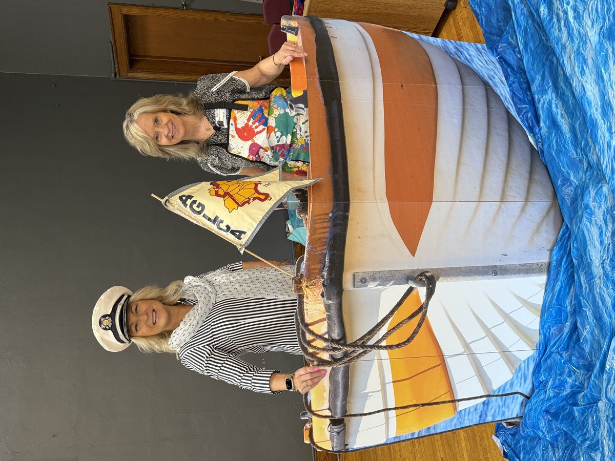 Kelly in a captain's hat and Jean in a colorful apron standing in the boat prop with the AGLCA flag