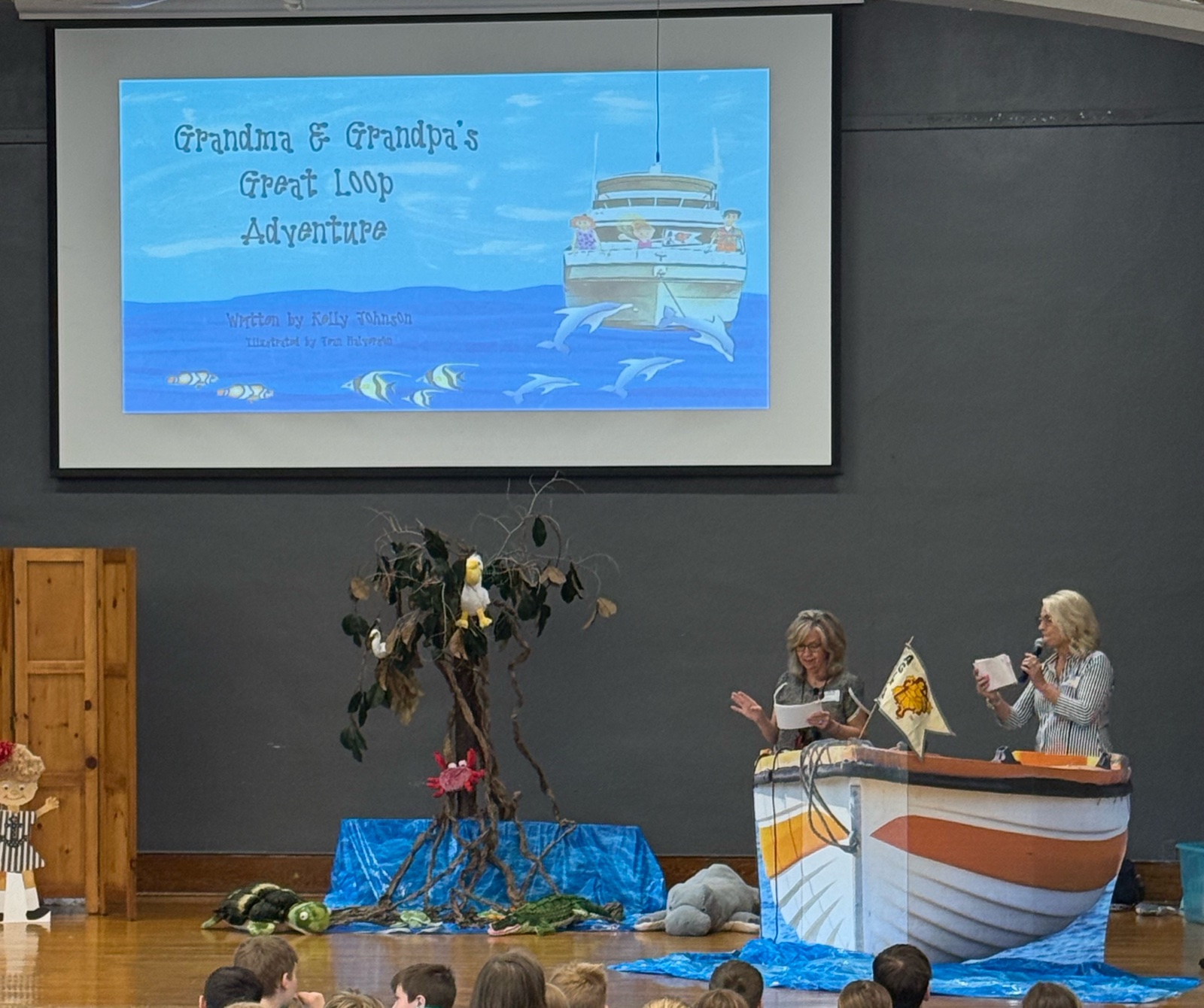 Kelly and Jean presenting at a school assembly with the boat prop, mangrove set, and book cover on screen